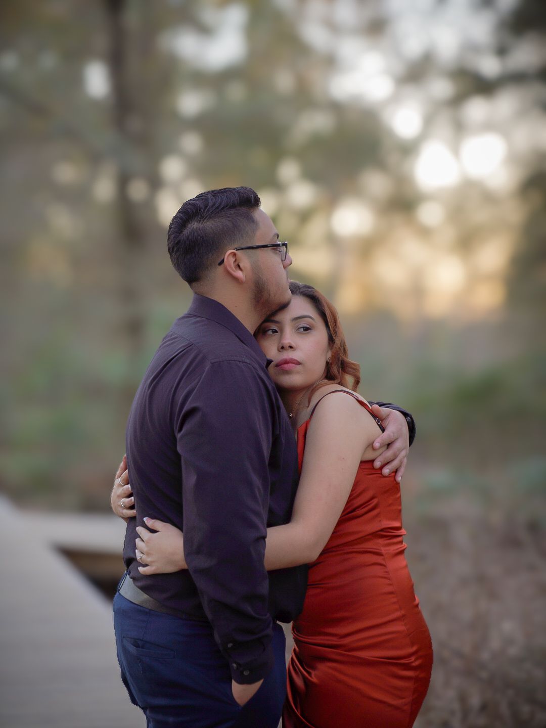 Couple embracing tenderly outdoors at dusk, woman in red dress leaning against man in purple shirt.