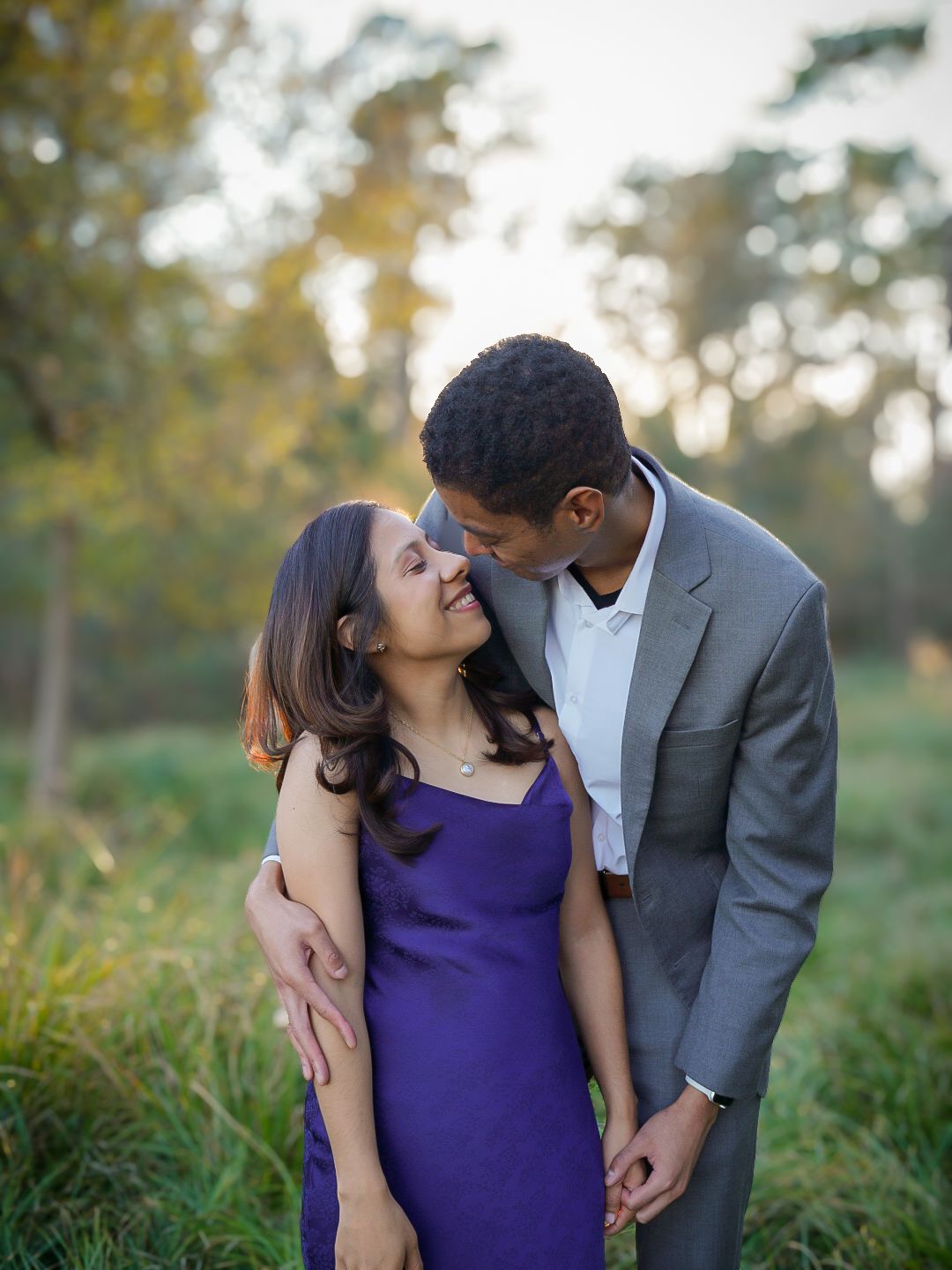 Couple sharing a tender moment in a lush green park, woman in purple dress, man in gray suit.