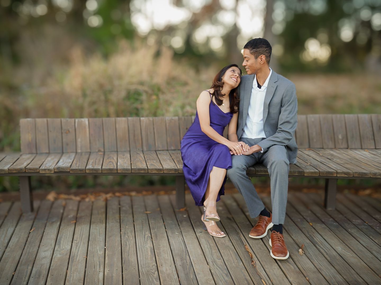 Couple sitting close on a wooden bench outdoors, woman in purple dress and man in gray suit holding hands.