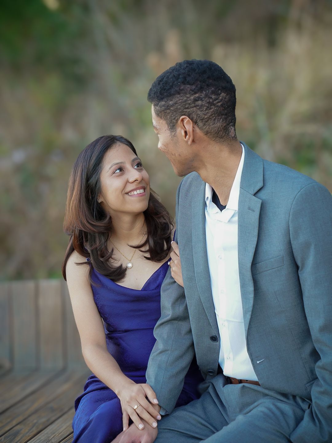 Couple sitting on wooden deck gazing at each other lovingly, woman in blue dress, man in gray suit.