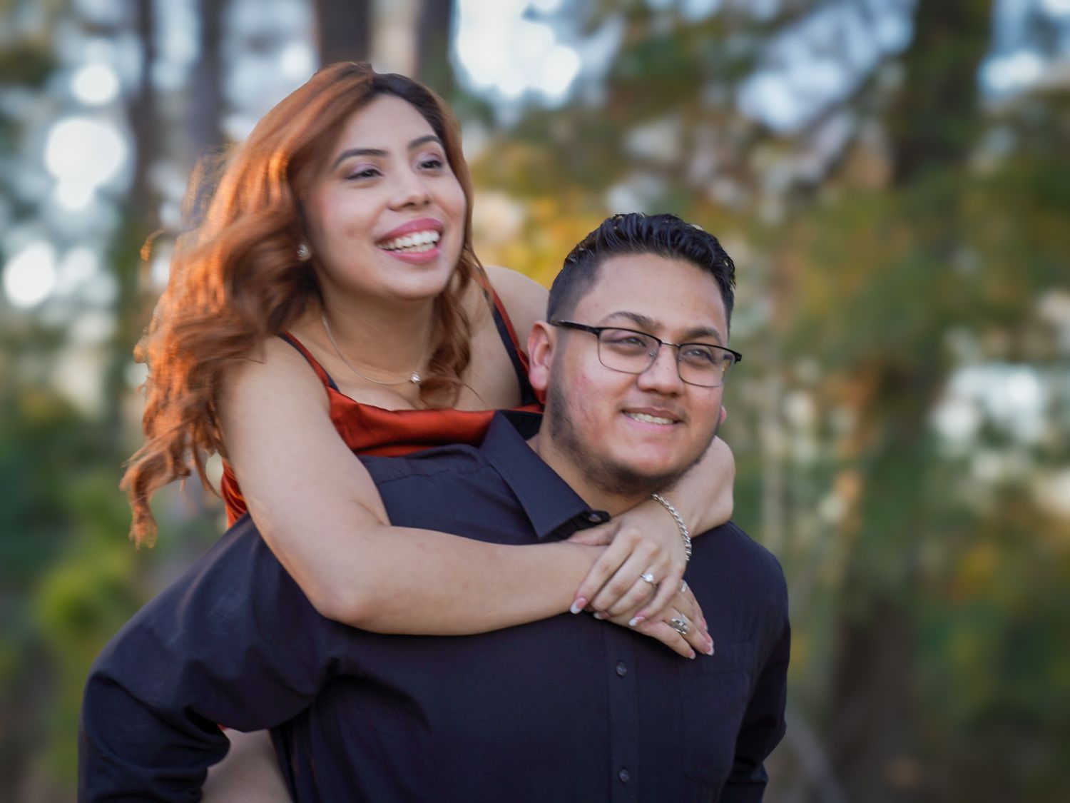 Woman with red hair piggyback riding smiling man in black shirt among autumn trees.