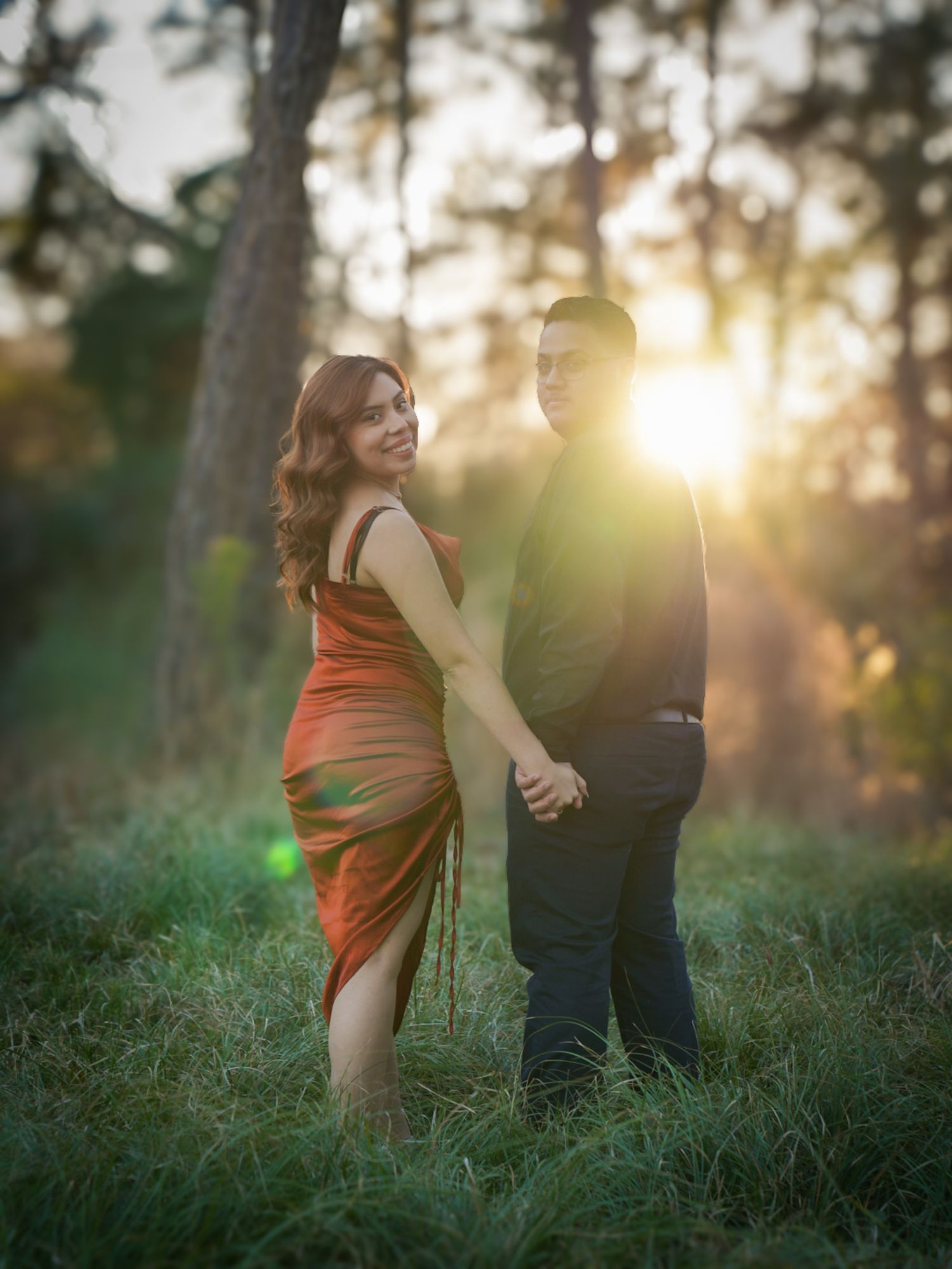 Couple holding hands at sunset in a forest, woman in orange dress smiling, golden backlight through pine trees.