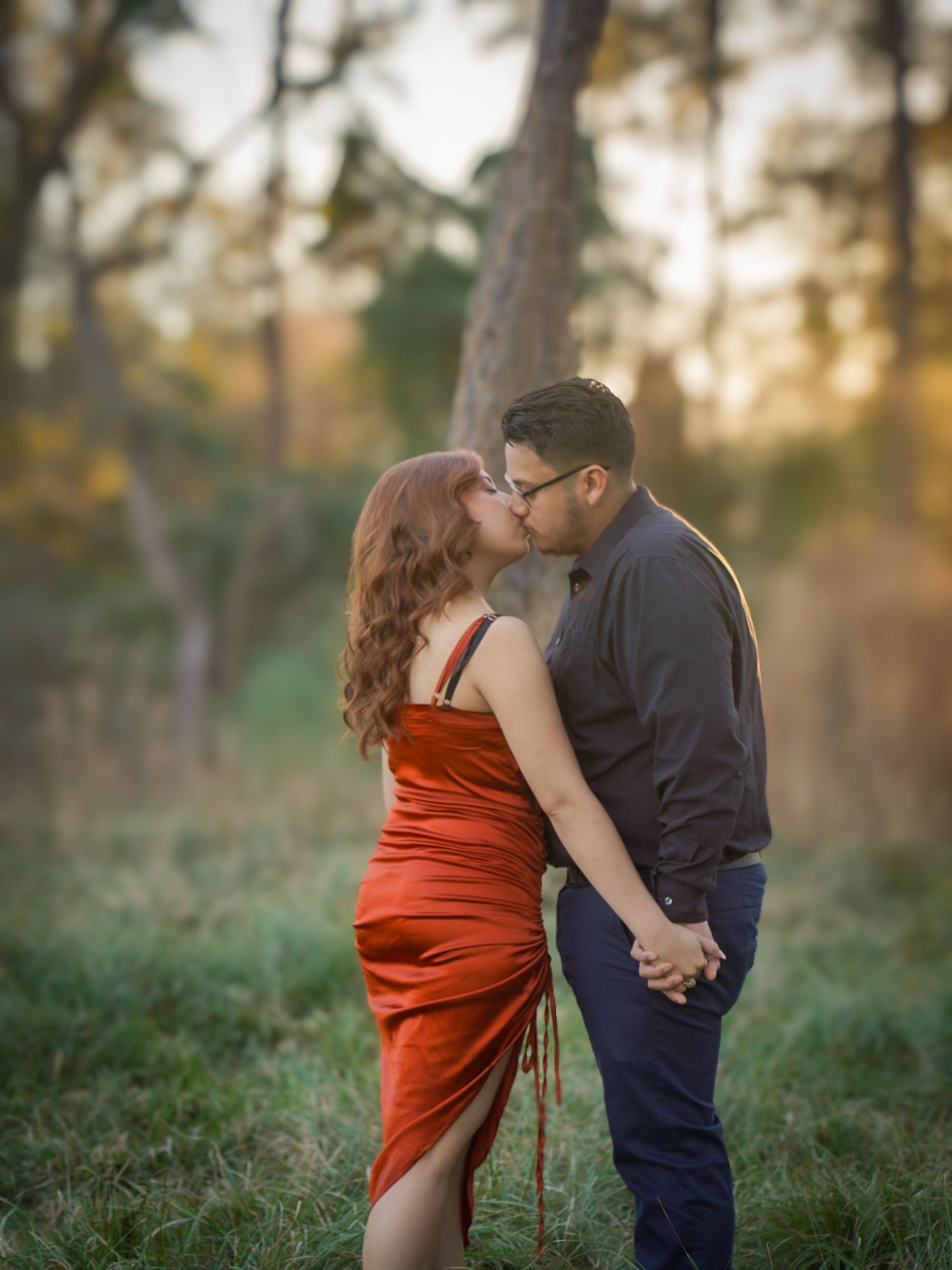 Couple sharing a kiss in a sunlit meadow, woman in red satin dress holding hands with man.