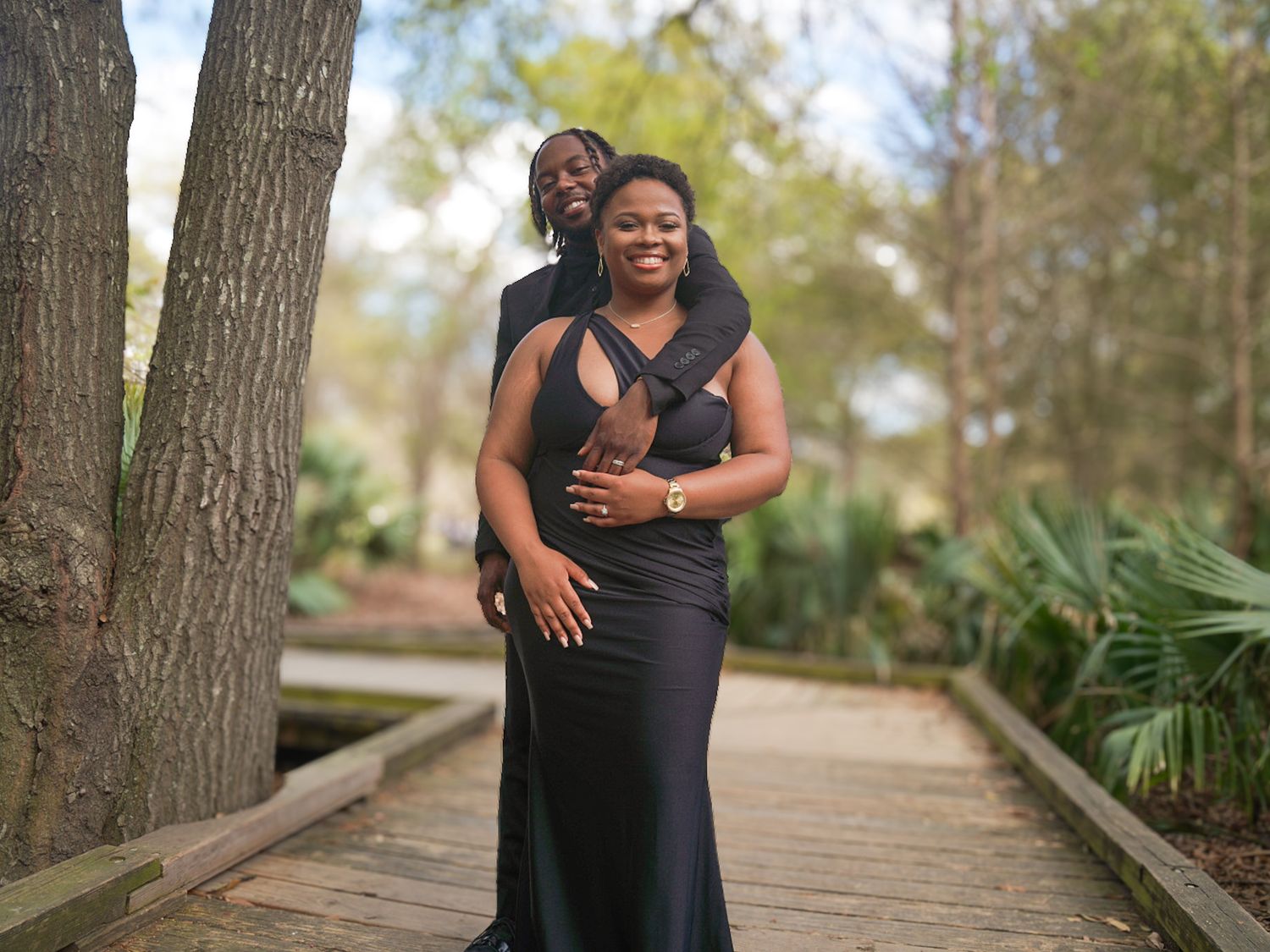 Couple posing on wooden boardwalk in park, woman in black gown, man standing behind her, surrounded by trees.