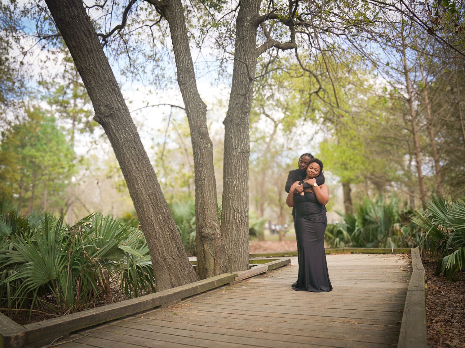 Couple embracing on a wooden boardwalk surrounded by trees in a scenic park setting.