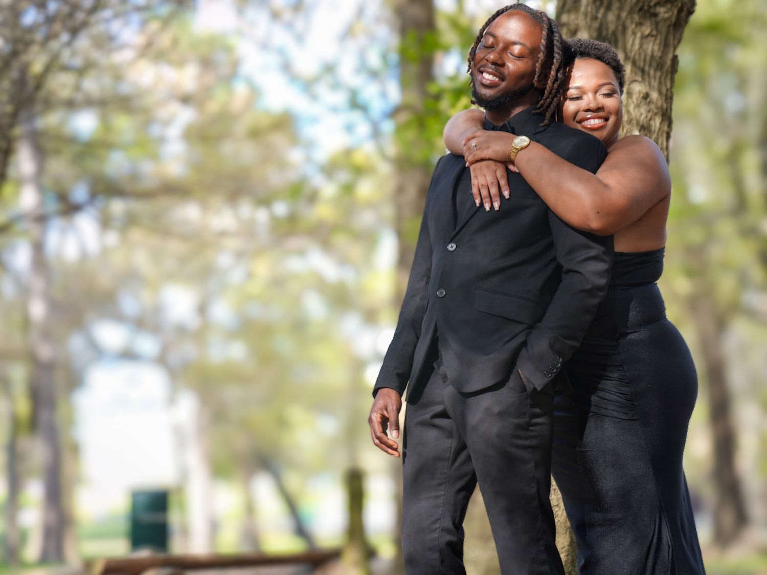 Couple in matching black formal attire sharing a joyful embrace in a sunny park setting.