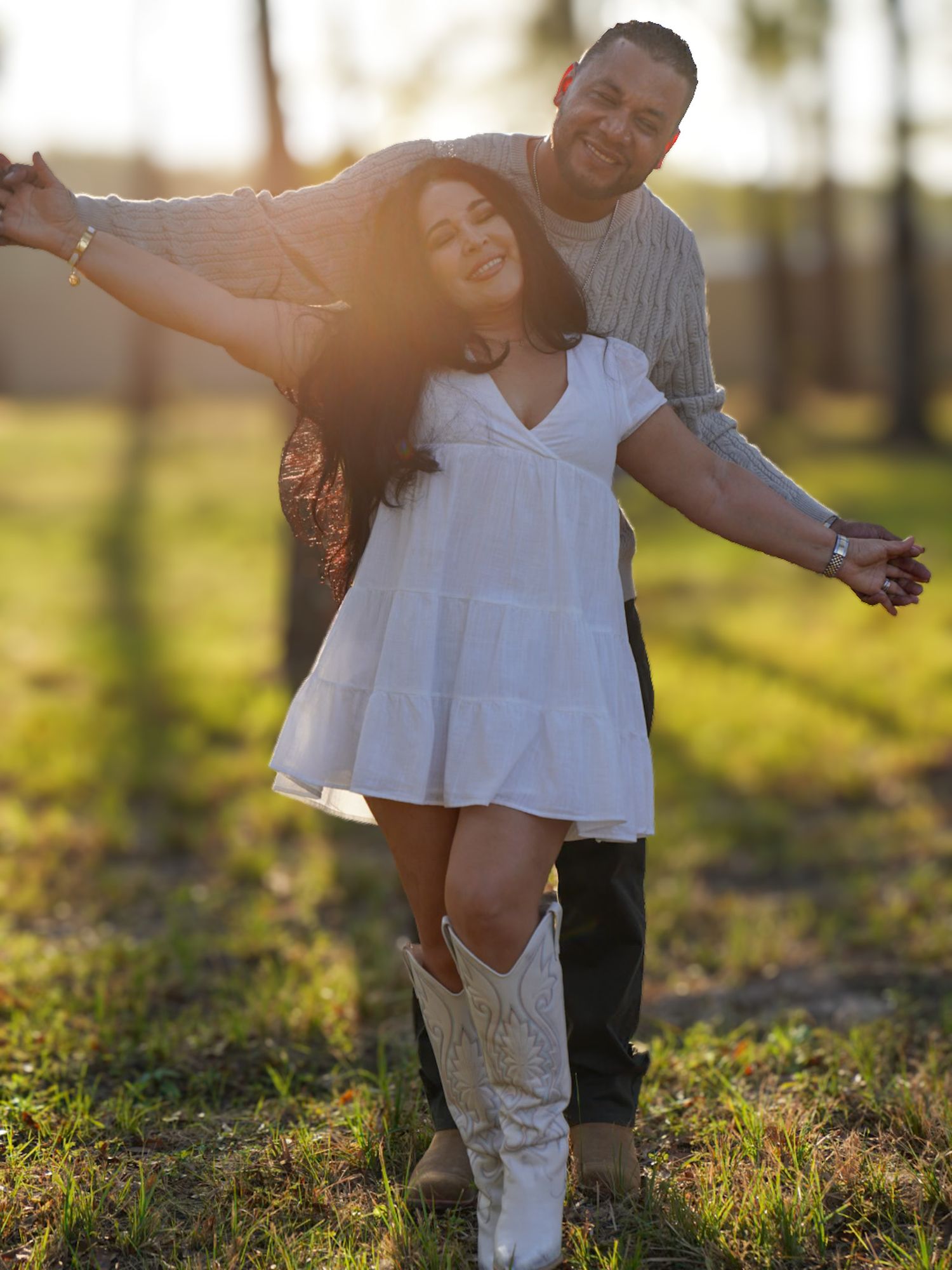 Couple dancing joyfully outdoors at golden hour, woman in white dress and cowboy boots, man embracing her from behind.