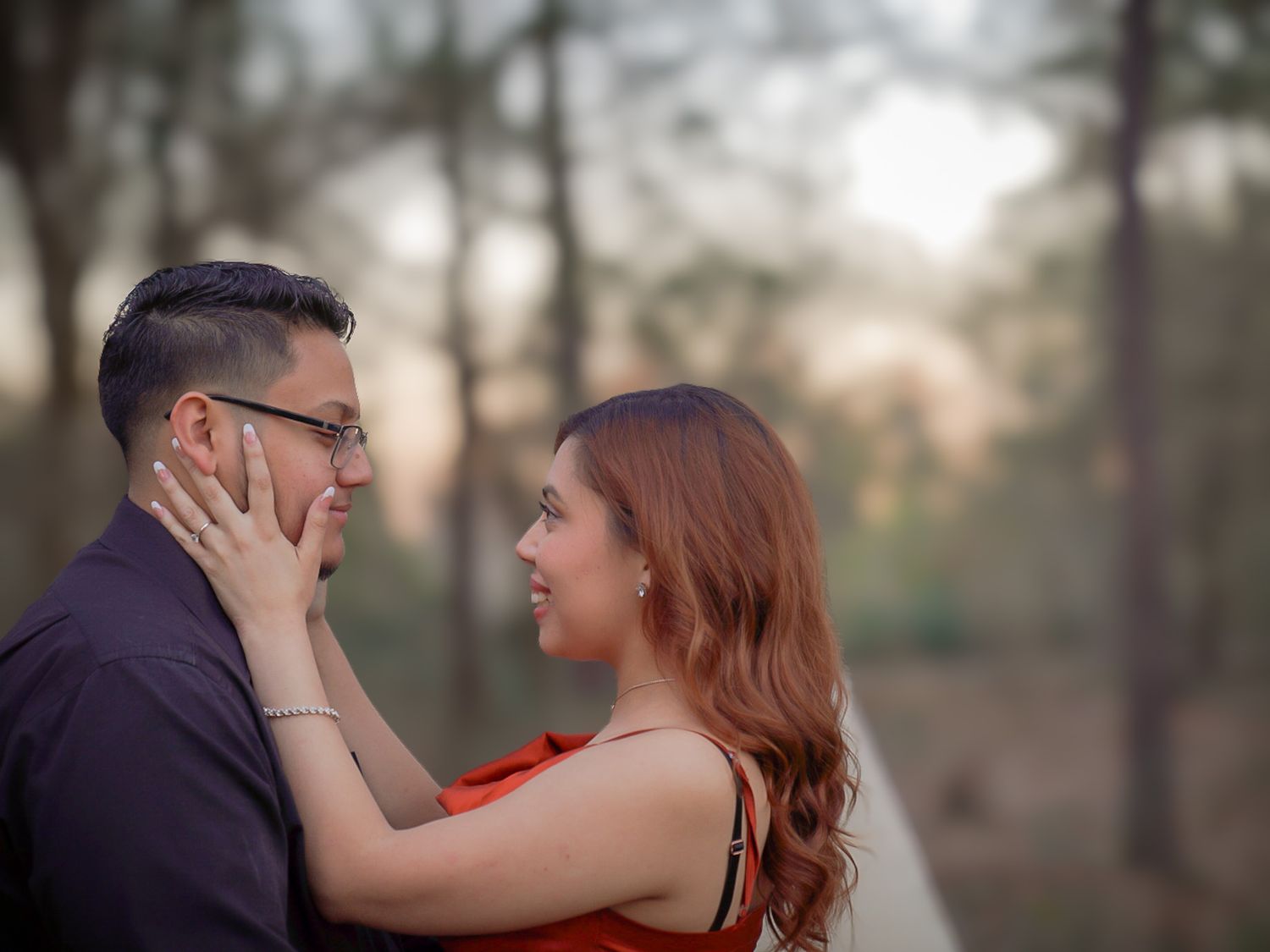 Romantic couple gazing into each others eyes in a sunlit forest, woman in red dress tenderly holding mans face.