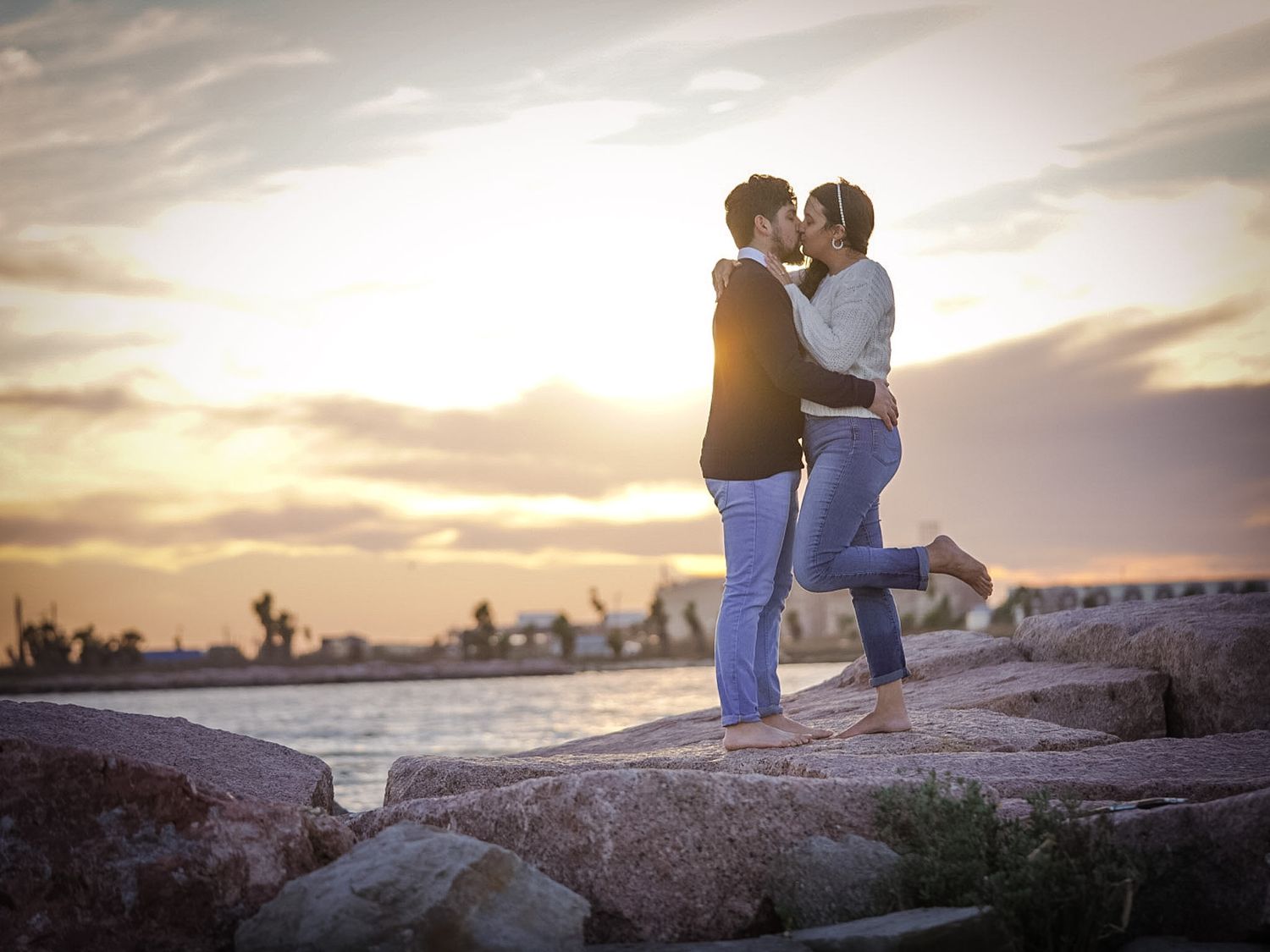 Romantic couple kissing on rocky shoreline during golden sunset by the water.