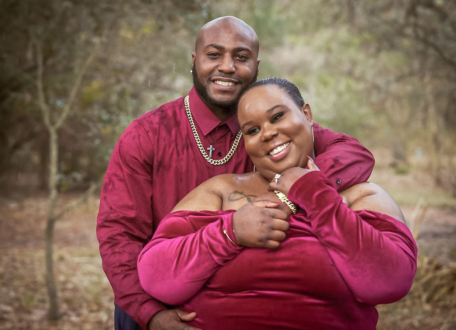 Couple in matching burgundy outfits embracing and smiling outdoors in a wooded setting.