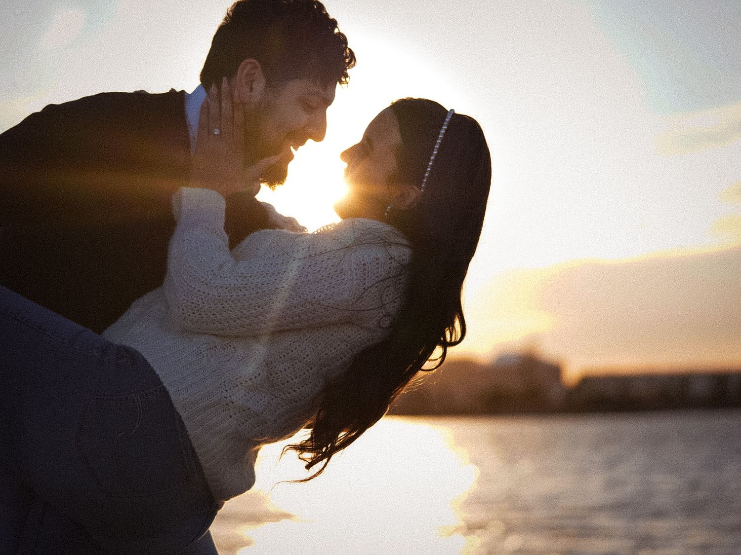 Romantic couple embracing at sunset by the water, golden light glowing behind them in a tender moment.