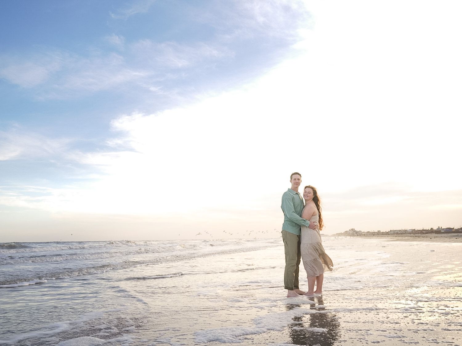 A couple stands together on a beach at sunset with waves at their feet.