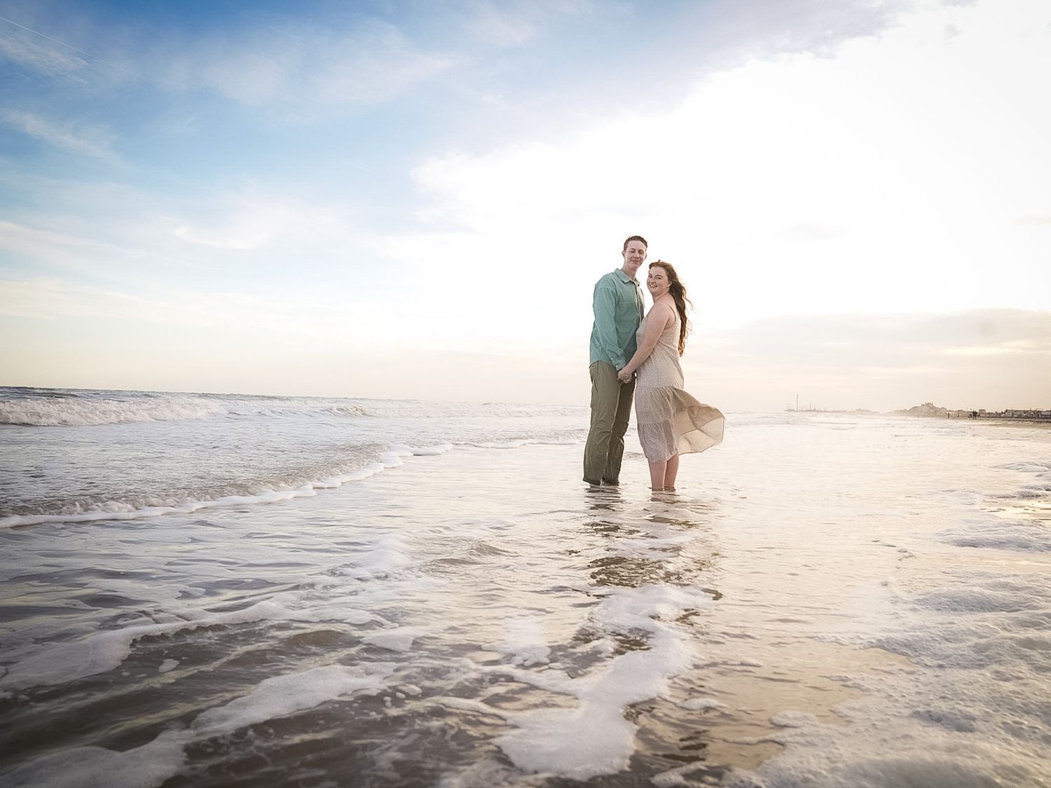 A smiling couple poses together on a sunlit beach with ocean waves nearby.