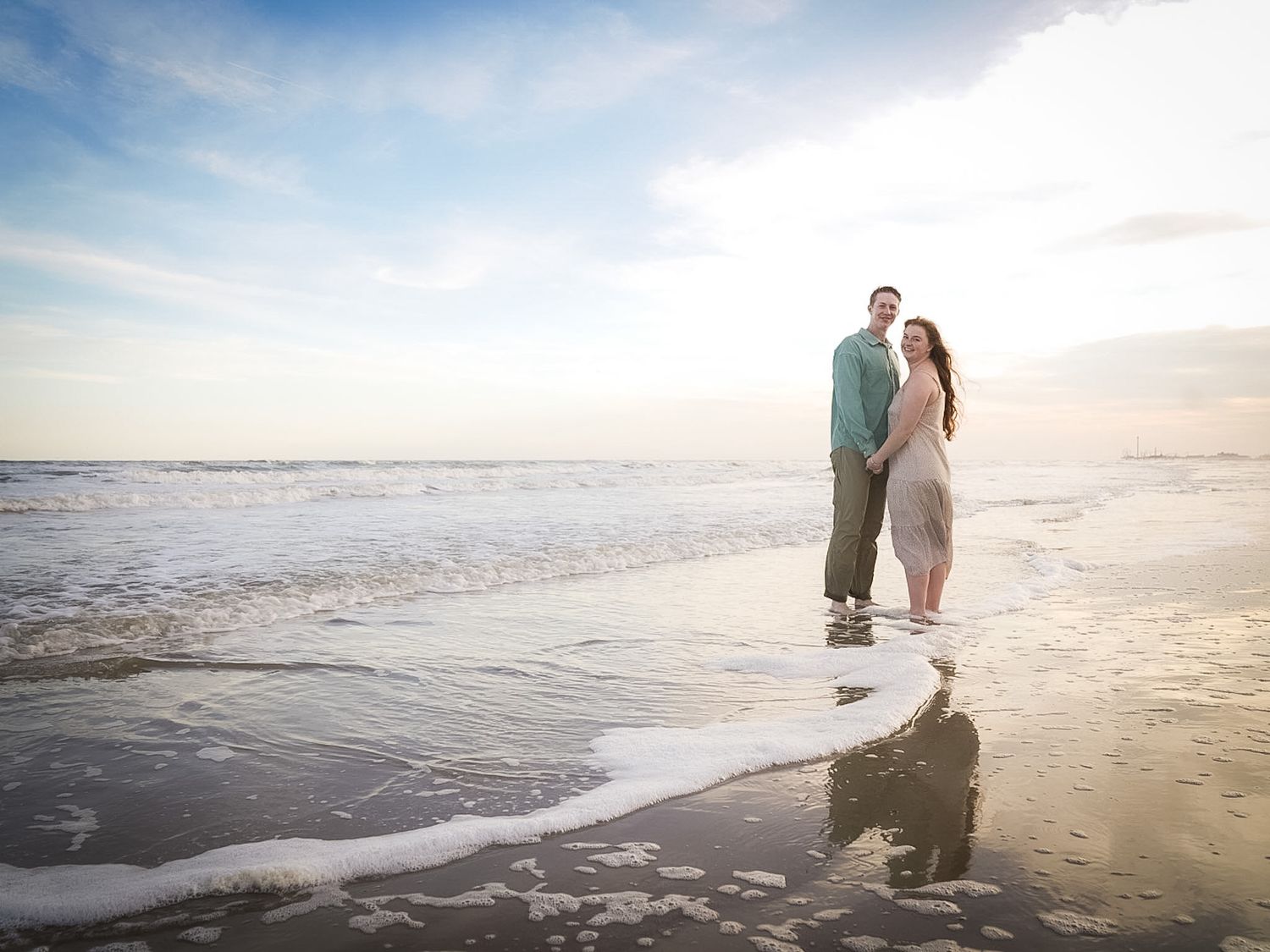Couple holding hands on a sunlit beach with ocean waves, reflecting on wet sand at golden hour.