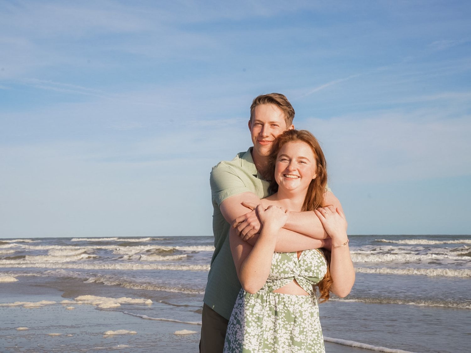 Happy couple embracing on a sunny beach with ocean waves, man hugging woman from behind, both smiling.