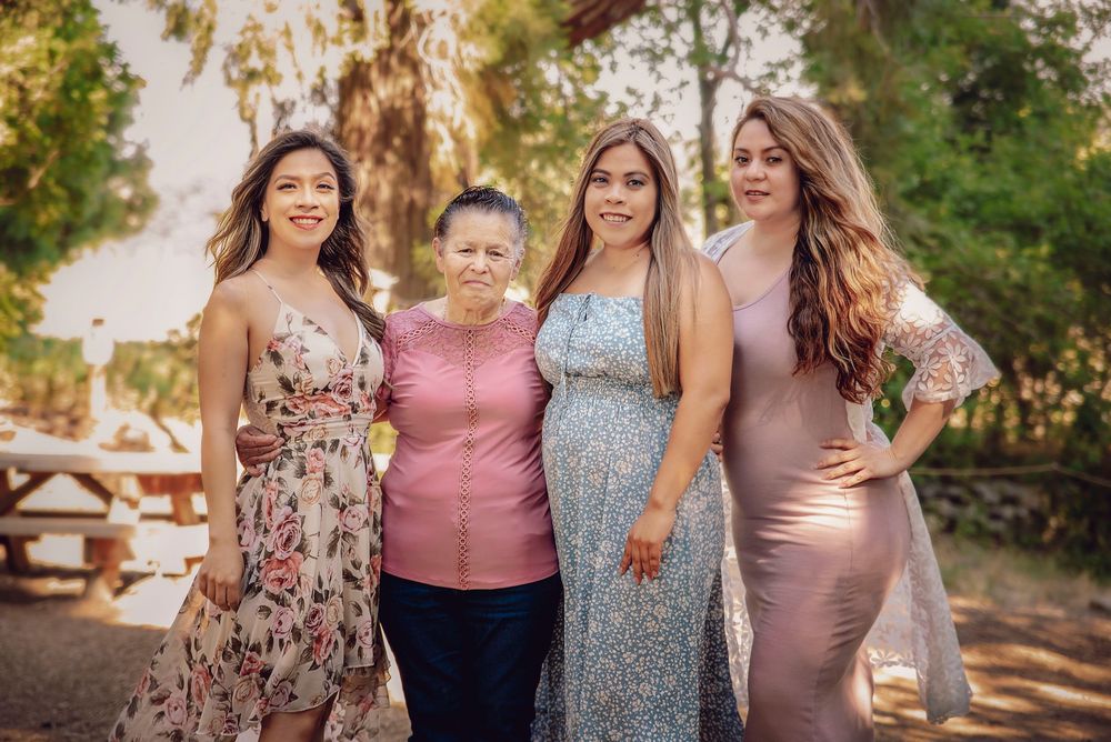 Four women in elegant dresses pose together outdoors at a family gathering surrounded by lush green trees.