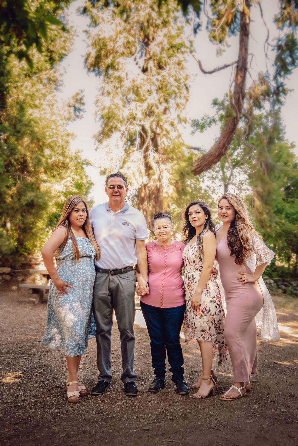 Family of five posing together outdoors on a dirt path surrounded by lush green trees during a warm sunny day.