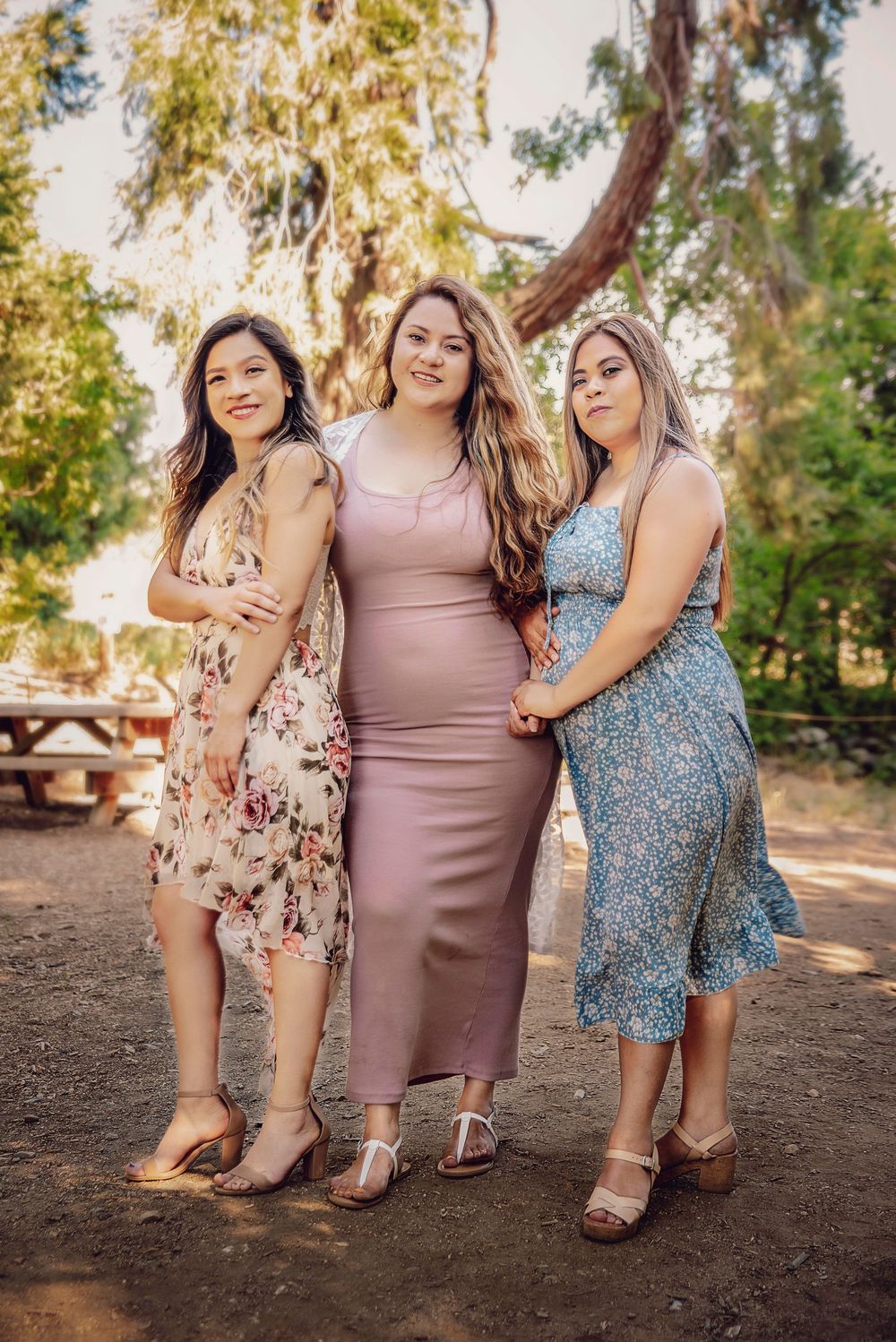 Three young women in summer dresses pose together outdoors among trees in a sunny park setting.