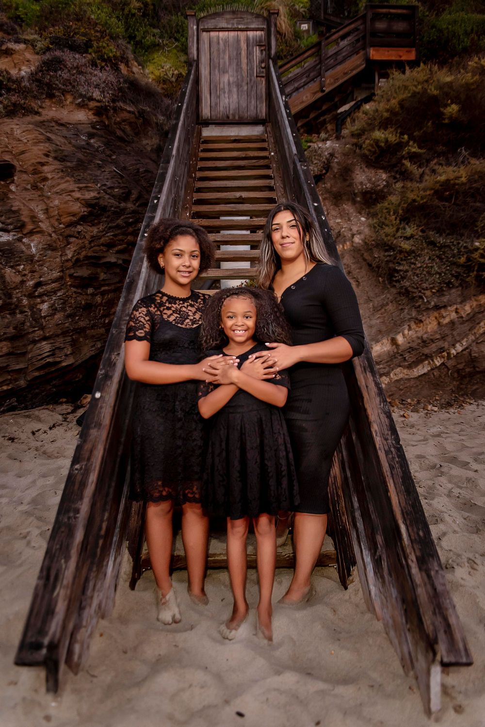 Three females in matching black dresses pose on a sandy beach in front of wooden stairs.