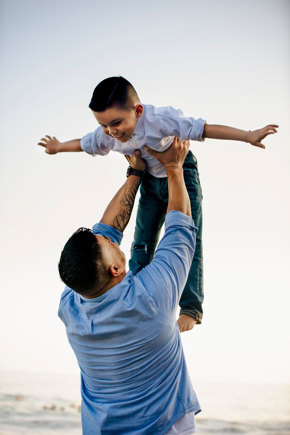 Father lifting young son in the air at the beach, child spreading arms like airplane, both smiling joyfully.