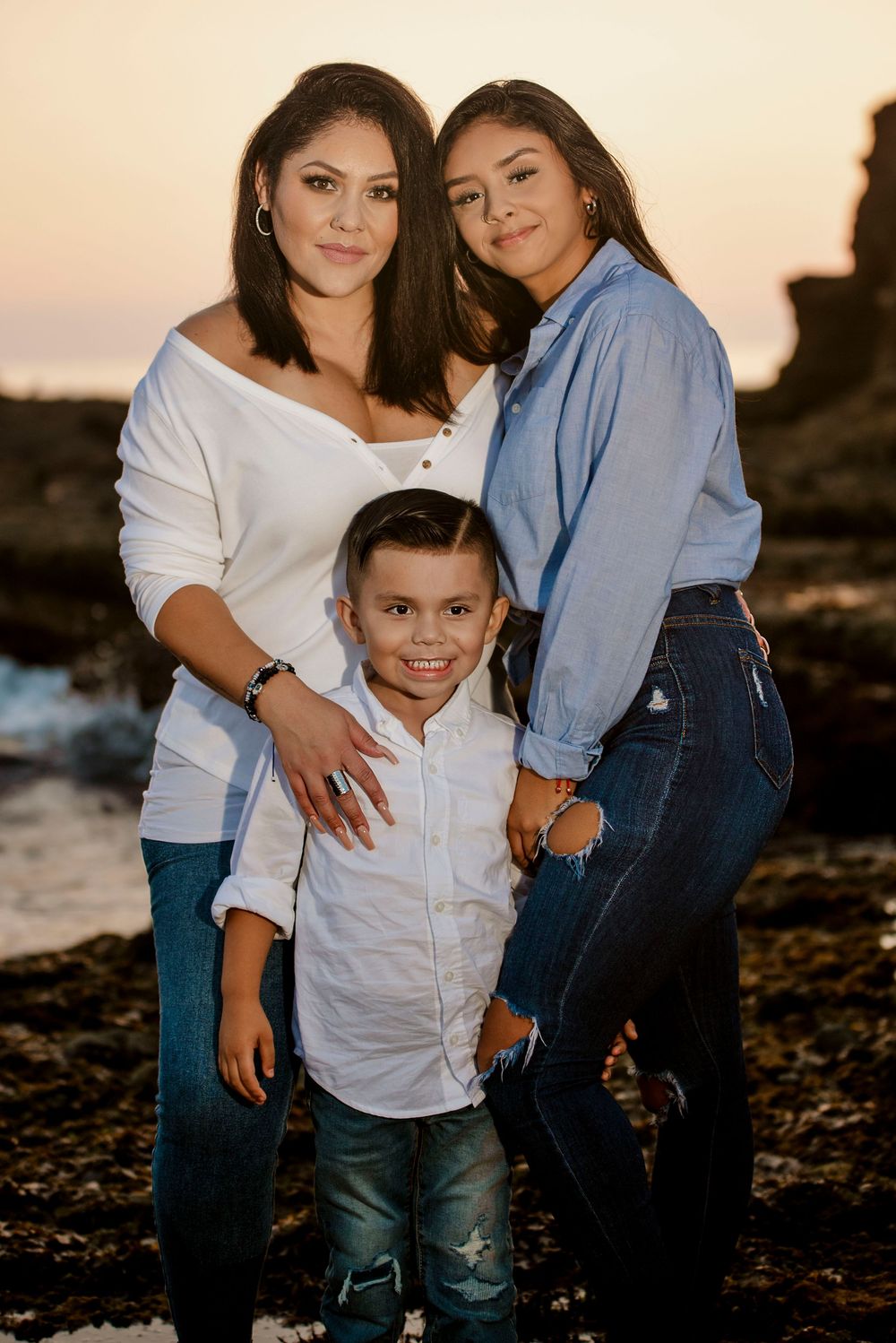 A woman and two children pose together on a rocky beach at sunset, wearing white and denim outfits.