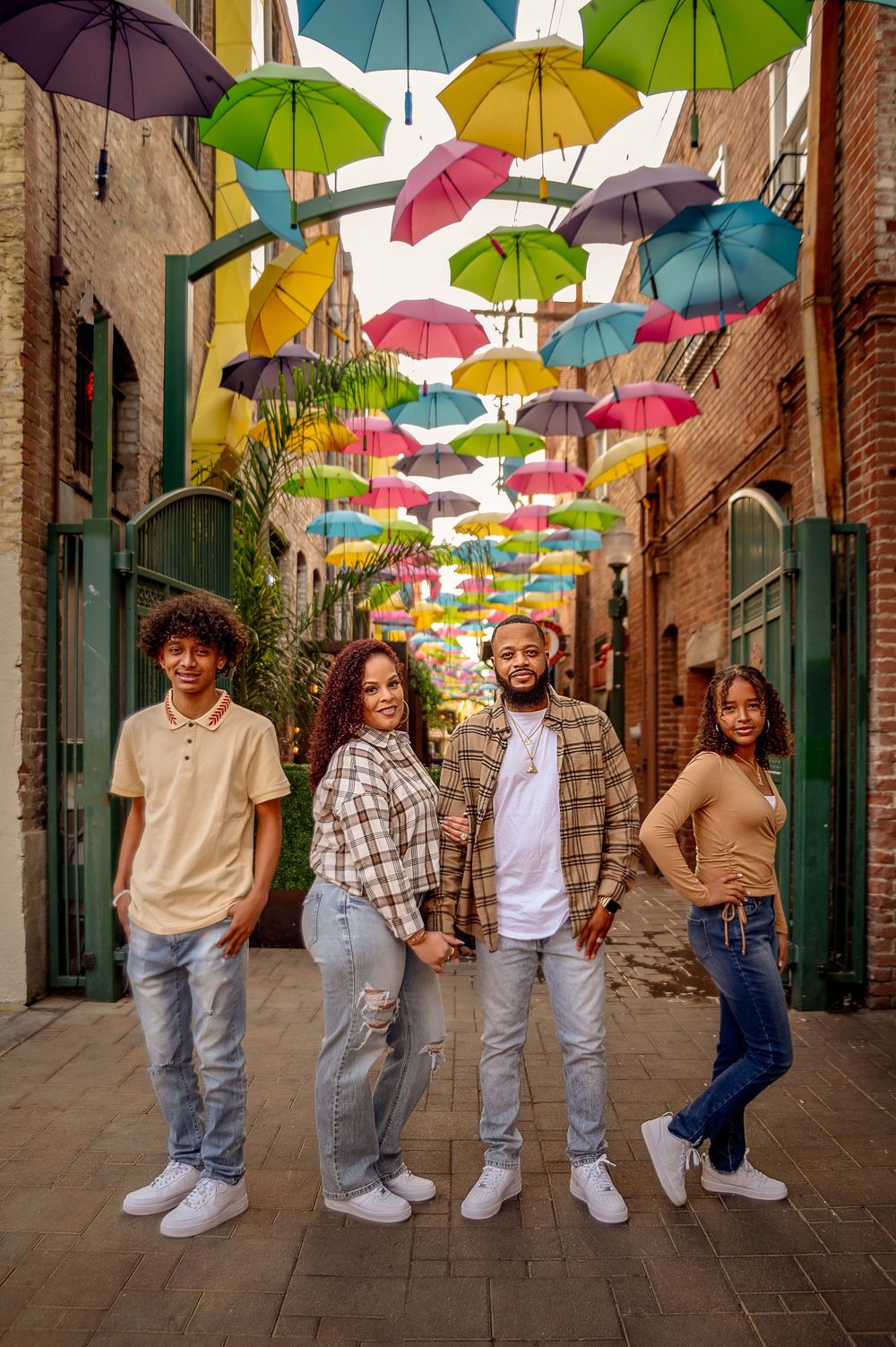 Four people pose in a colorful alley decorated with hanging multicolored umbrellas overhead.