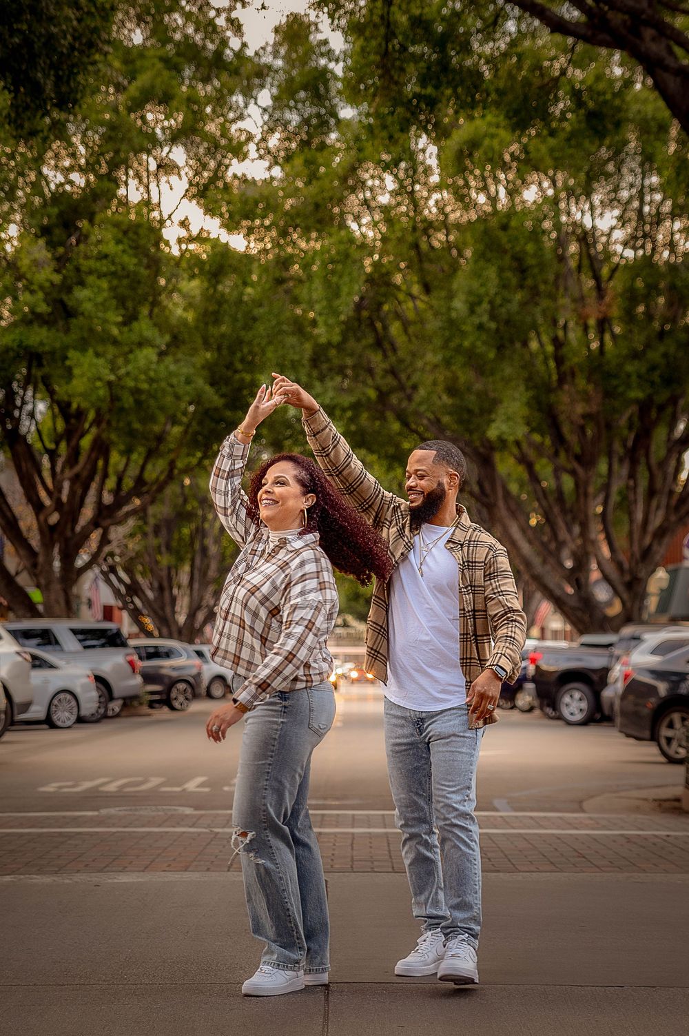 A joyful couple dancing outdoors in matching plaid shirts and jeans, surrounded by lush green trees in a parking lot.