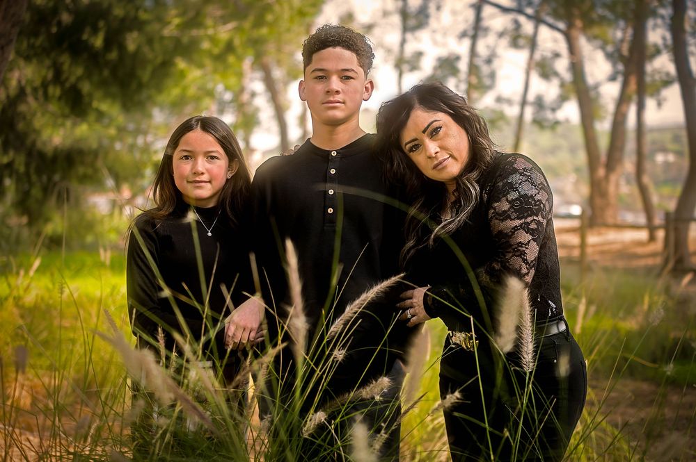 Three young people in black outfits pose together outdoors among tall grass with soft natural lighting.