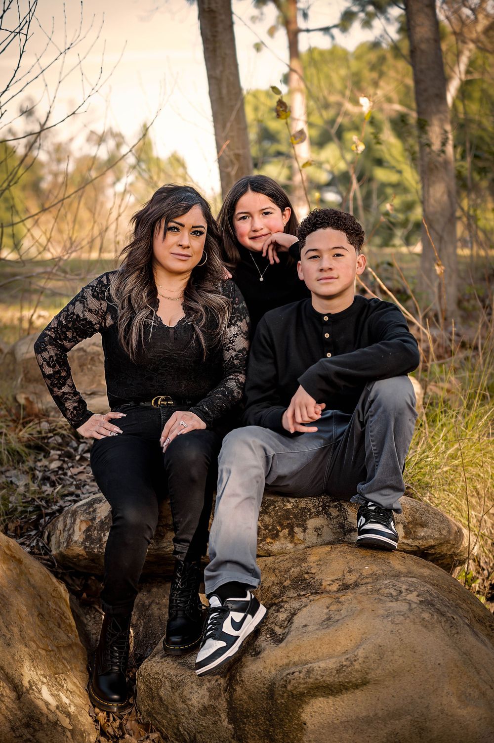 Family of three posing on rocks in a wooded outdoor setting, wearing coordinated black outfits during golden hour.