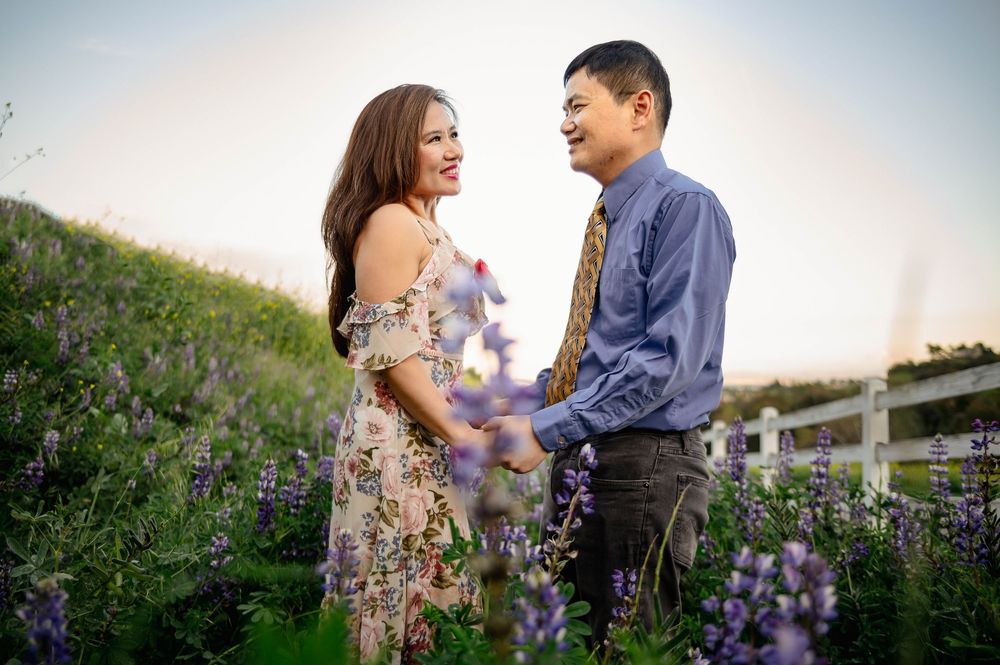 Couple smiling at each other in a field of purple wildflowers at sunset, woman in floral dress, man in blue shirt.