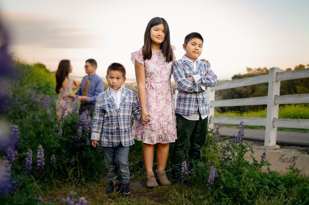 Three children pose outdoors near a white fence surrounded by purple wildflowers at golden hour.