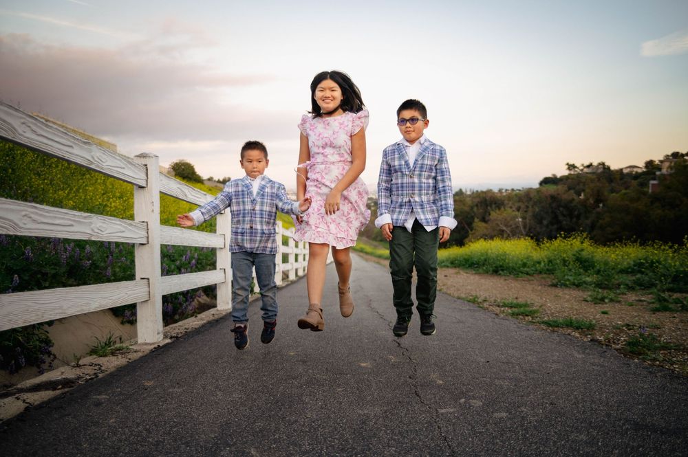 A woman and two boys jump joyfully on a path near a white fence with green hills in the background.