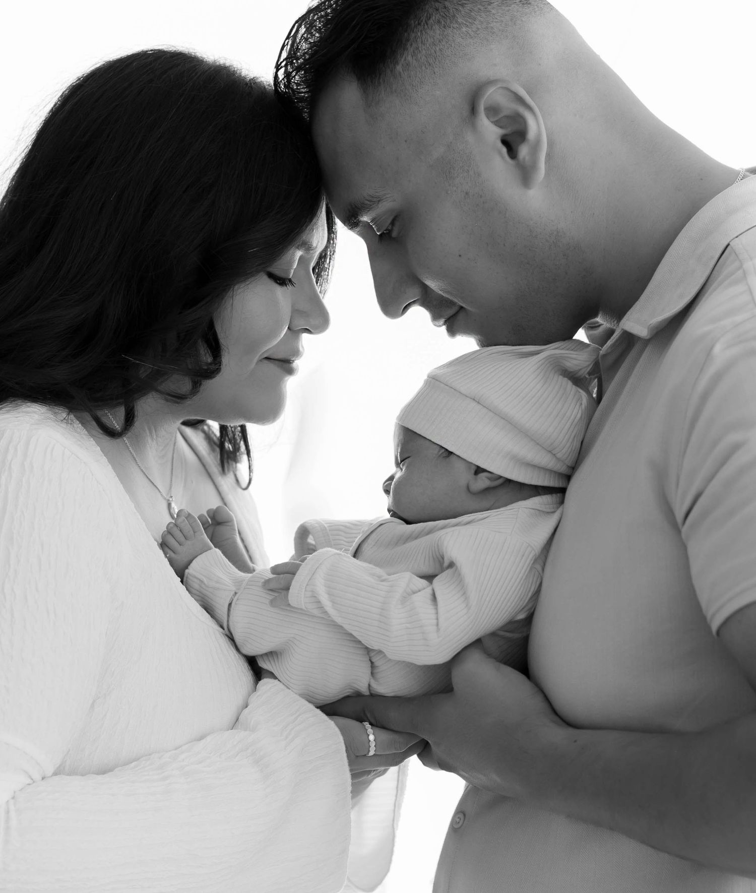 New parents in white outfits lovingly hold their newborn baby together against a bright white background.