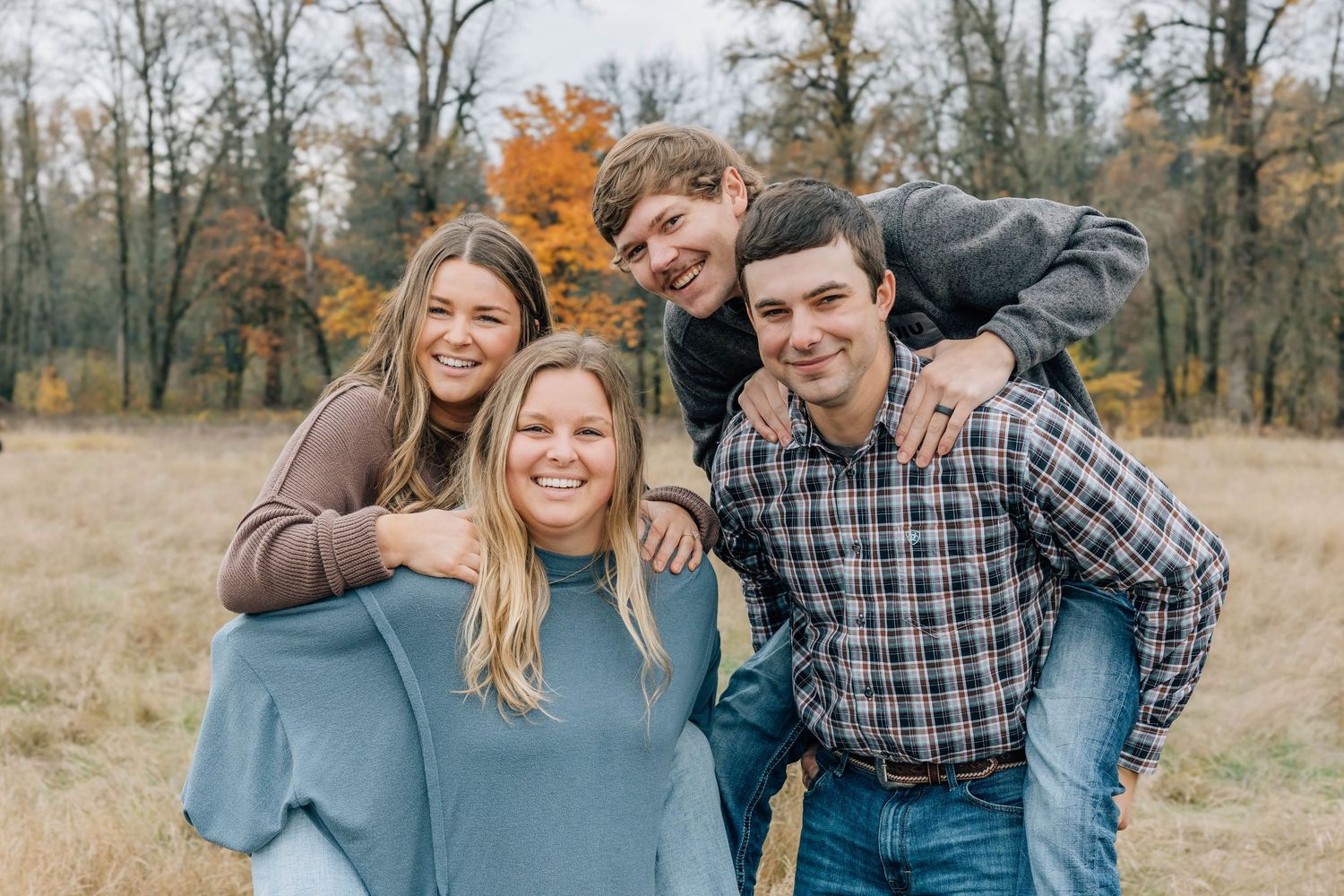 Happy family of four posing outdoors in autumn field with colorful fall foliage trees in background.