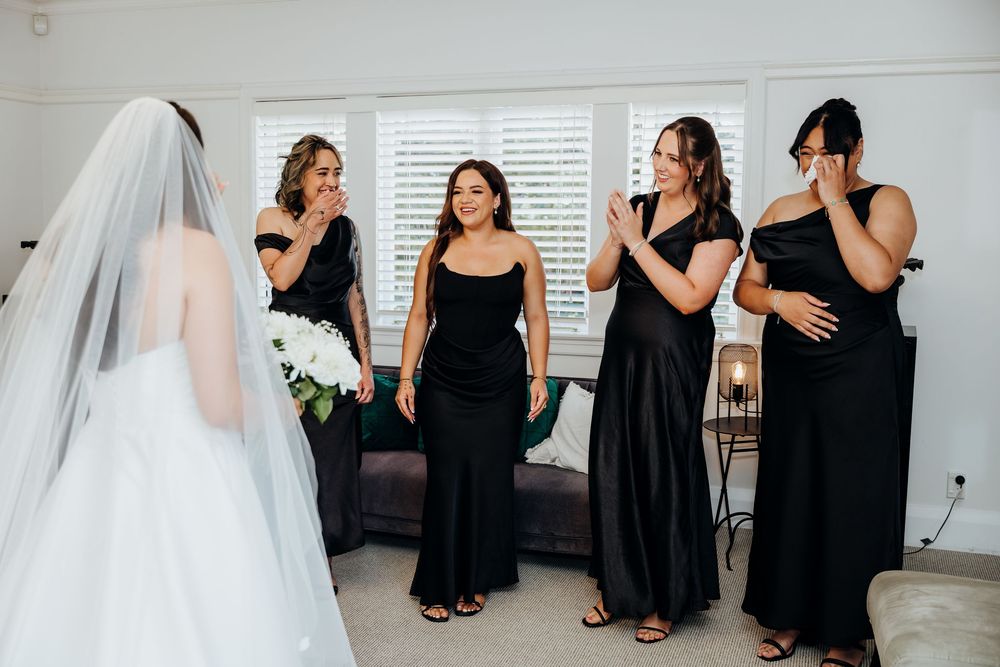 Bride in white gown with veil showing her look to emotional bridesmaids in black dresses.