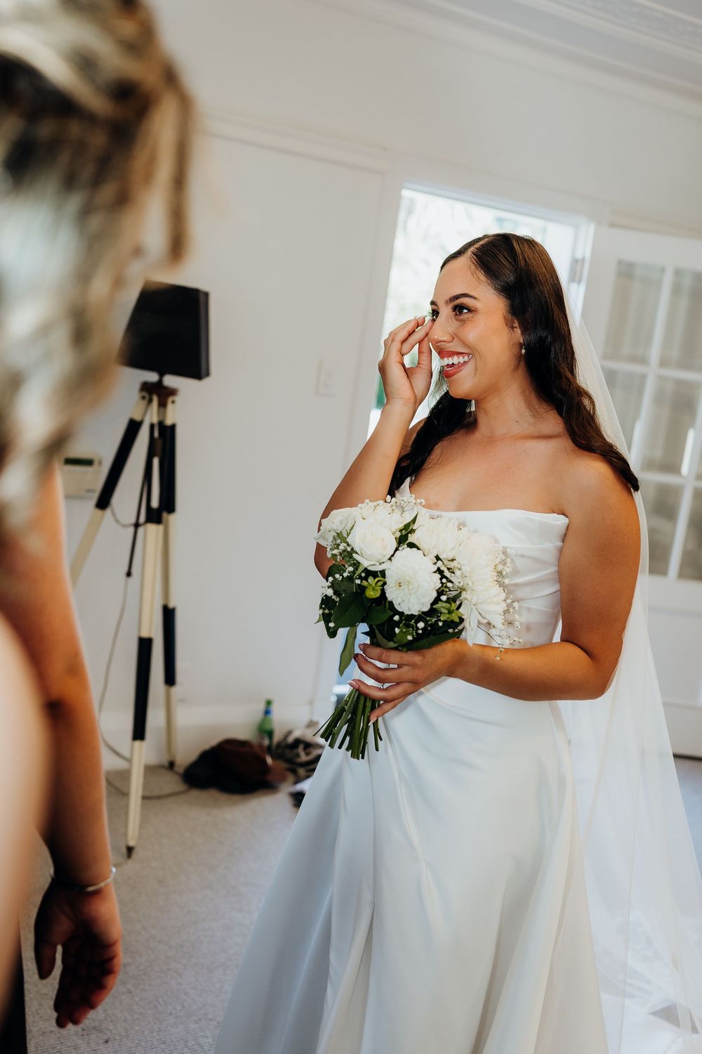 Emotional bride in strapless white gown holds white floral bouquet while wiping tear during wedding preparations.