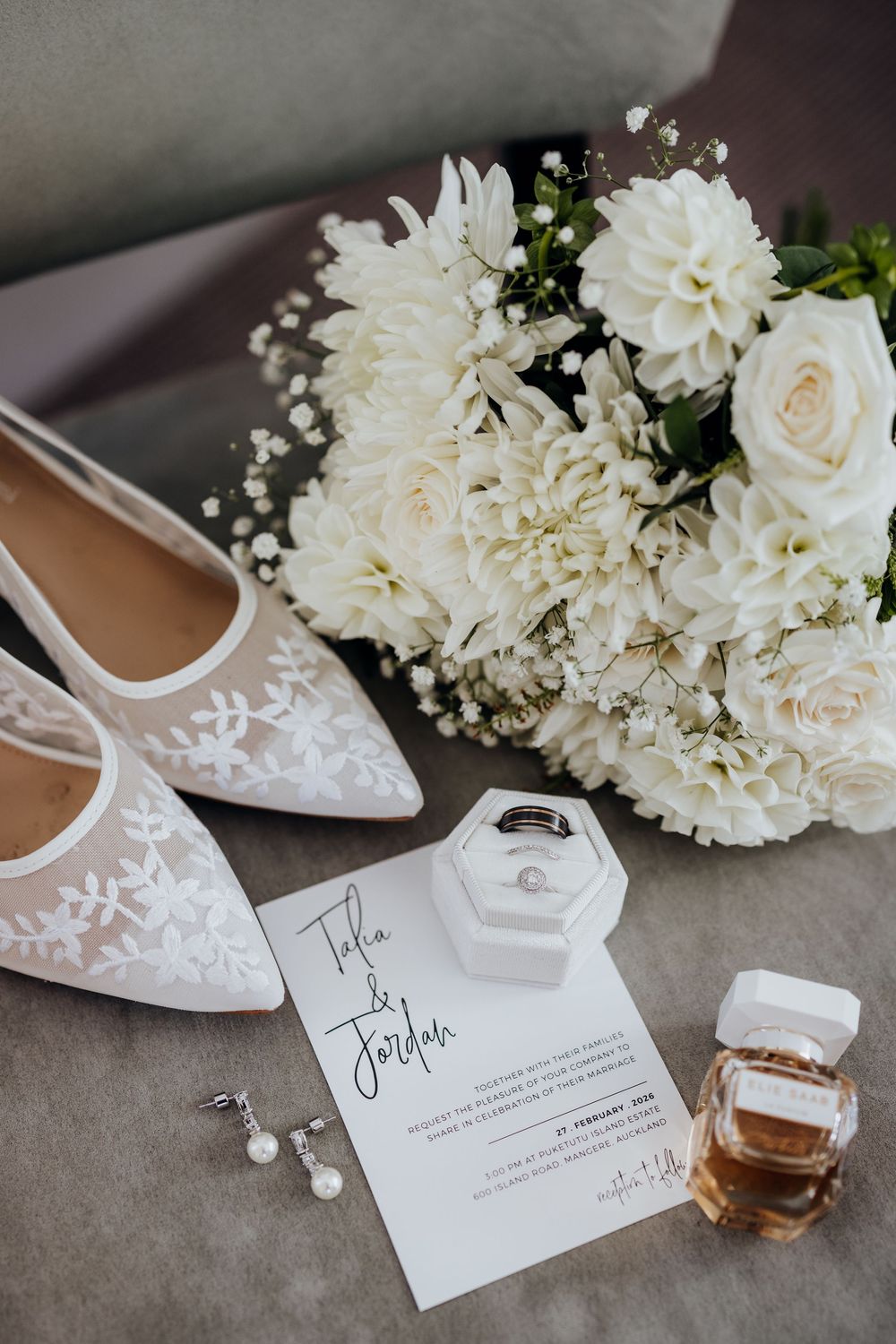 Elegant wedding flat lay featuring white bridal heels, floral bouquet, rings, invitation, earrings, and perfume.