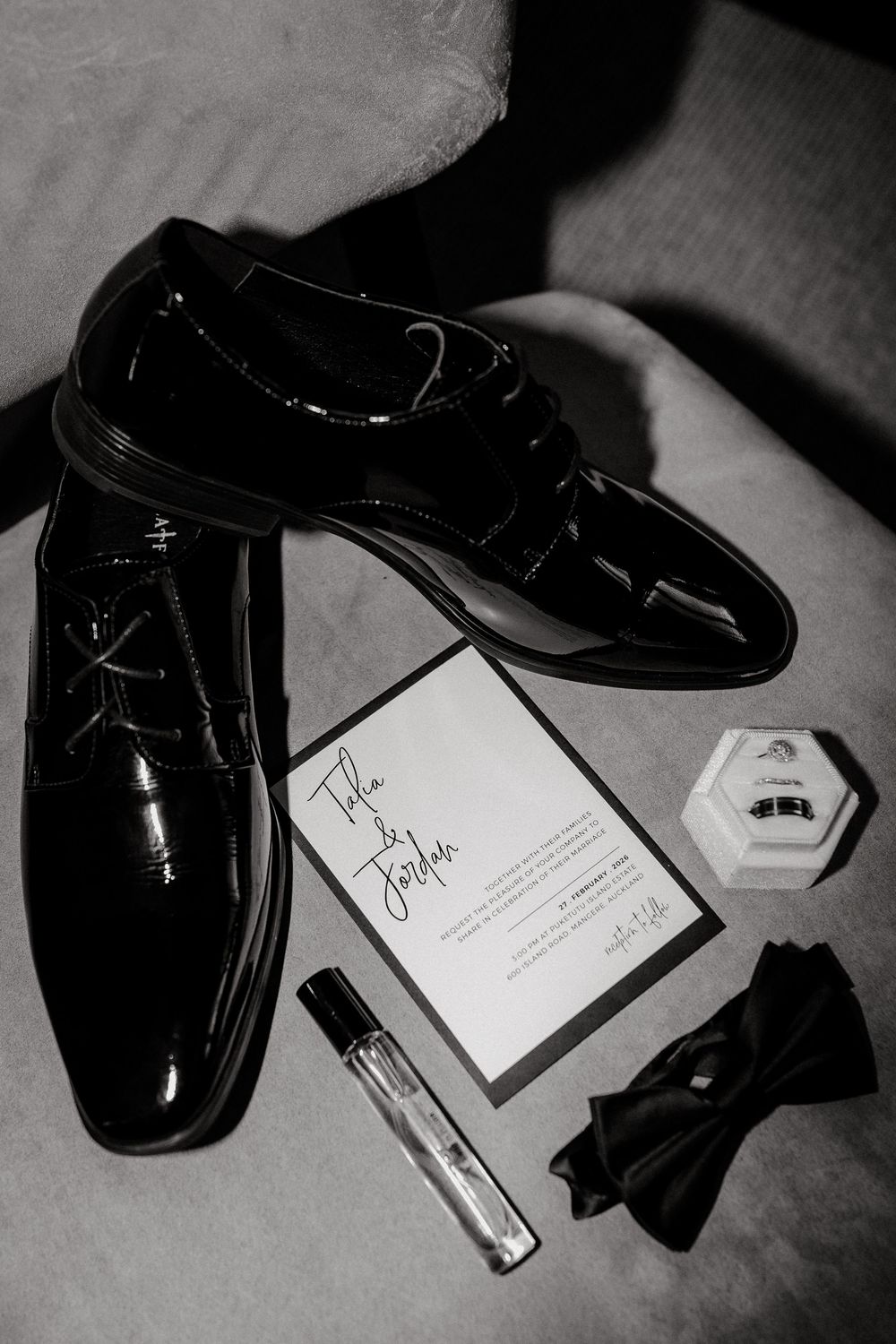 Black and white flat lay of groom wedding accessories including patent leather dress shoes, invitation, cologne, ring box, and bow tie.