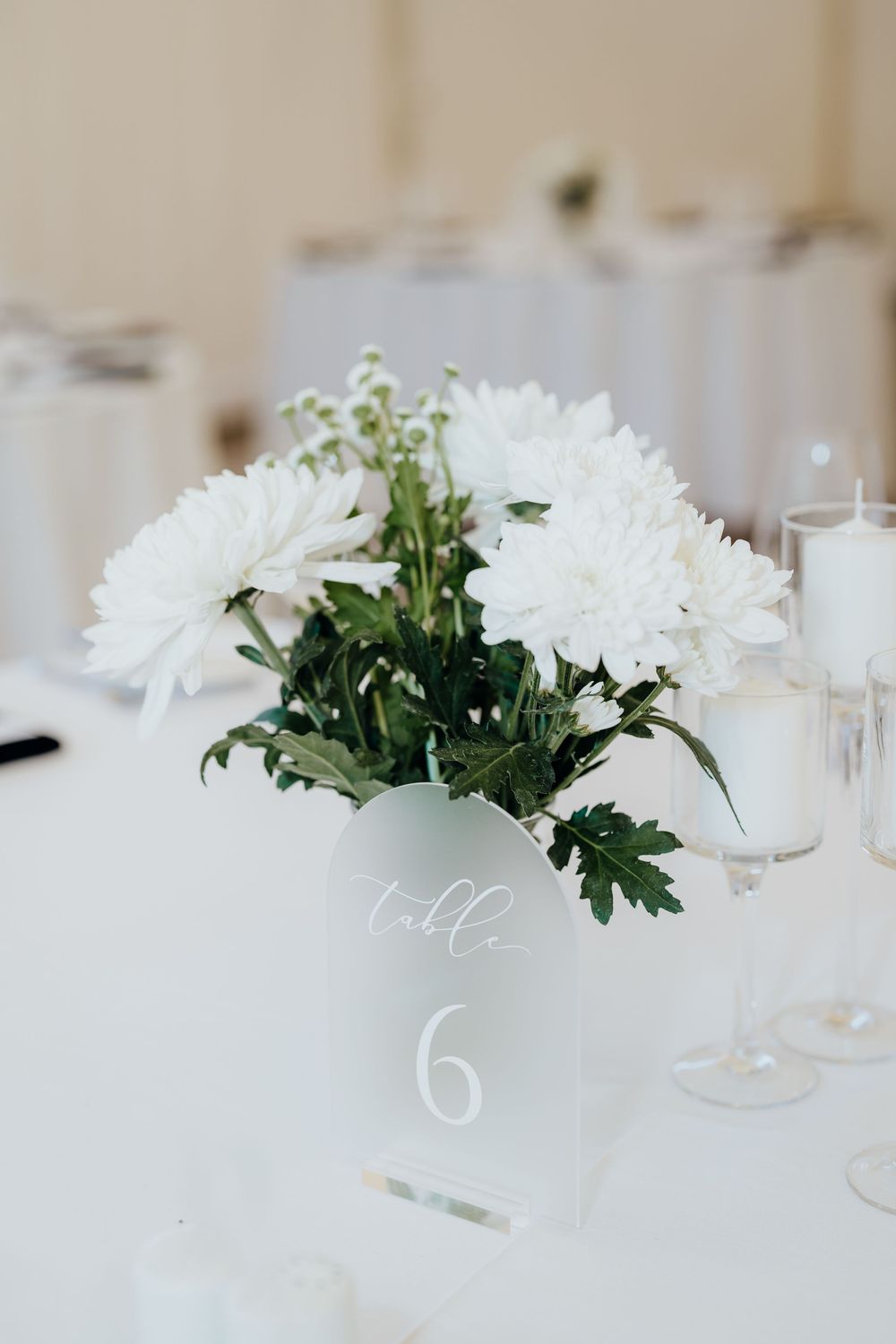 White chrysanthemum wedding centerpiece in white vase labeled Table 6 with candles on white tablecloth.