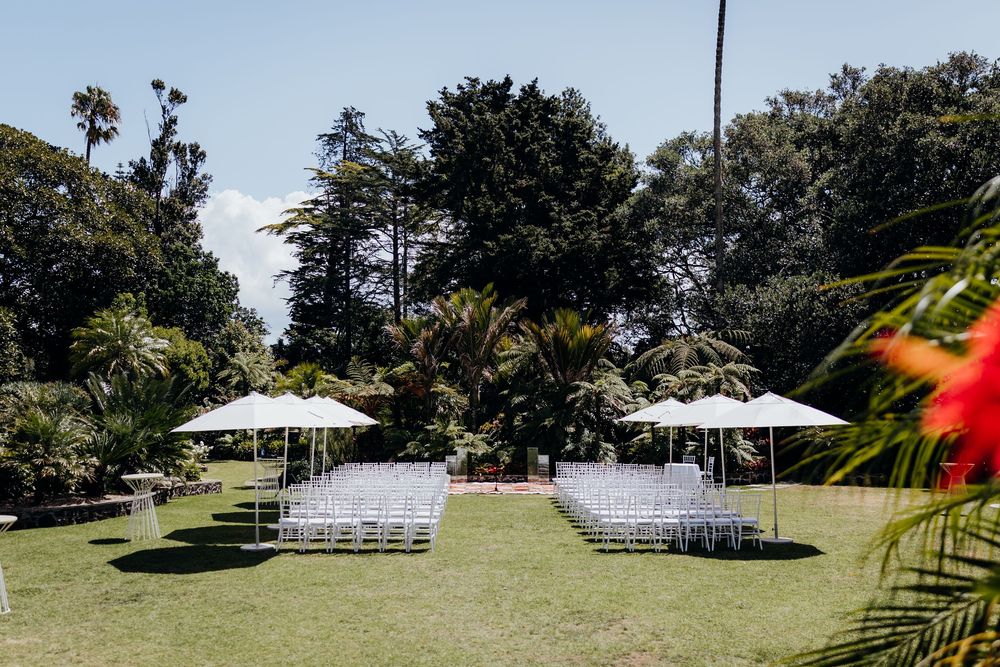 Outdoor wedding ceremony setup with white chairs and large umbrellas on lush green lawn surrounded by tropical trees.