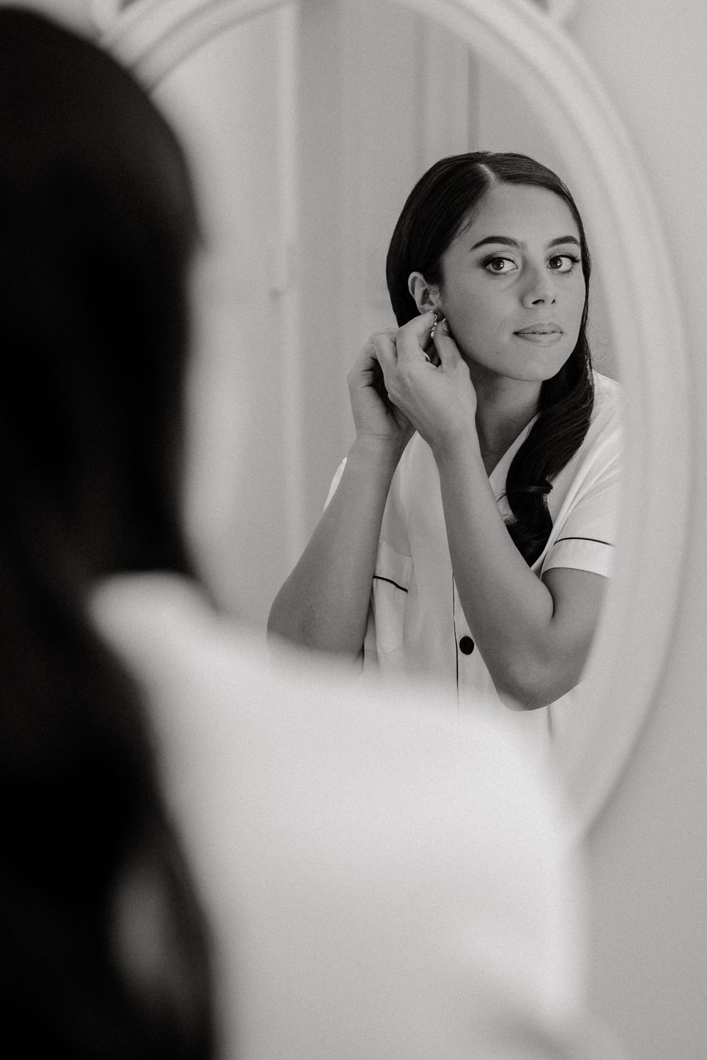 Black and white photo of bride putting on earrings while looking in oval mirror during wedding preparations.