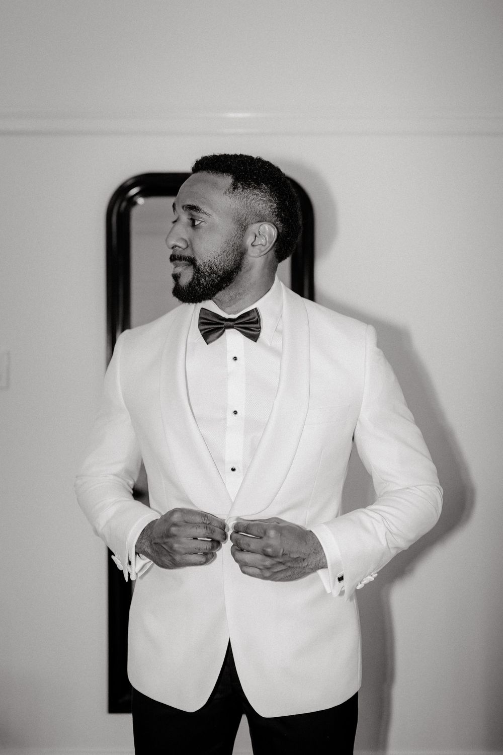 Black and white photo of a groom in a white tuxedo jacket and bow tie buttoning his jacket before his wedding.