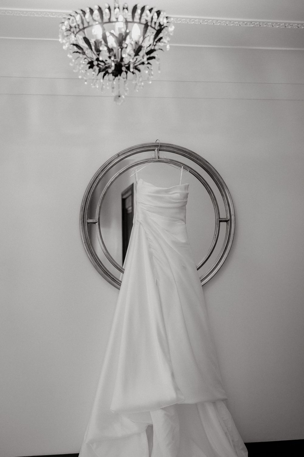 Elegant white wedding dress hanging on a round mirror with a crystal chandelier above in black and white.