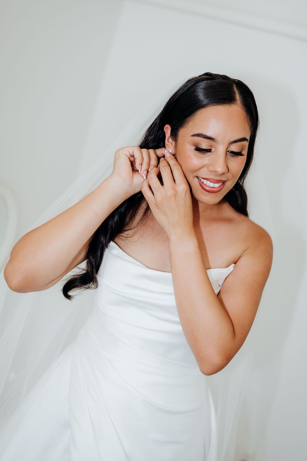 Smiling bride in a strapless white gown adjusts her earring while getting ready on her wedding day.