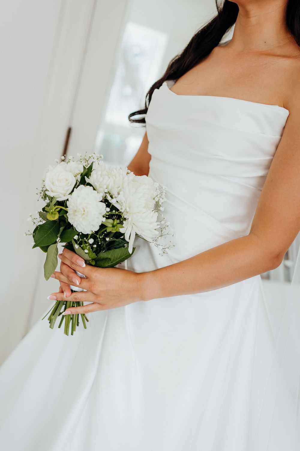Bride in strapless white wedding gown holds a delicate bouquet of white roses and greenery.