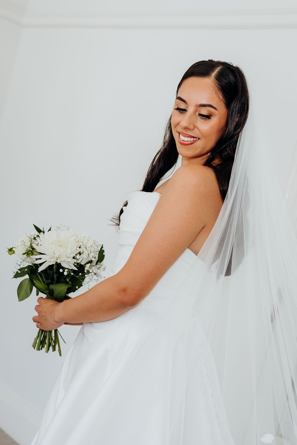 Radiant bride in a strapless white gown and veil holds a white floral bouquet, smiling softly.