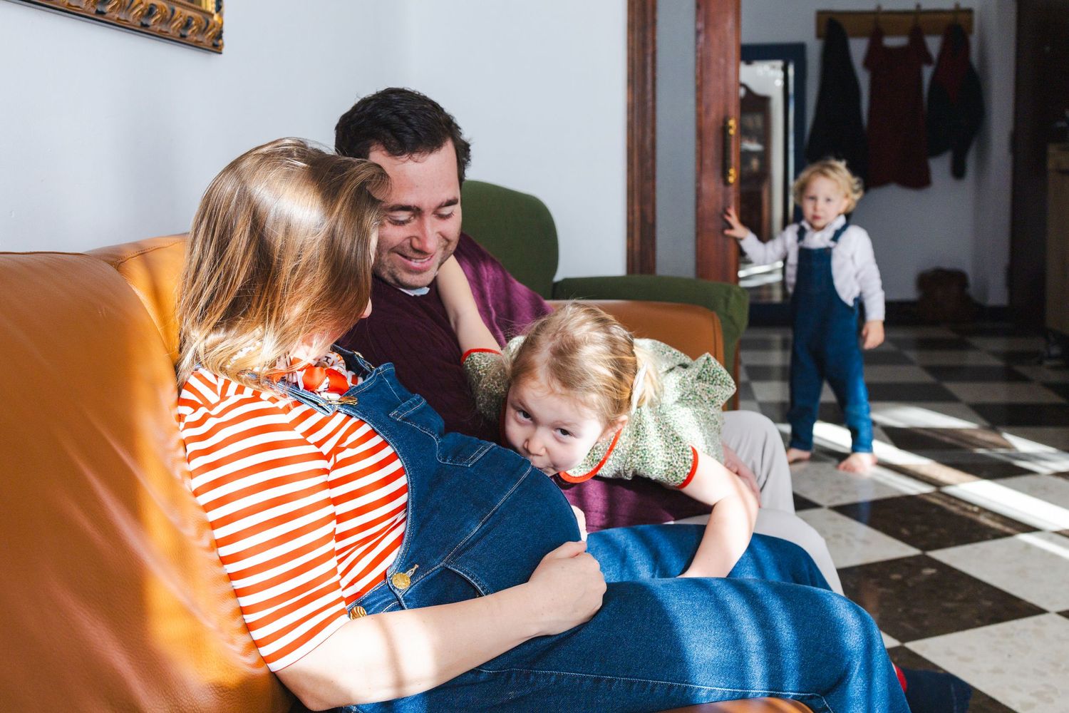 Pregnant woman and man sit on sofa smiling at toddler while a child dances in background on checkered floor.