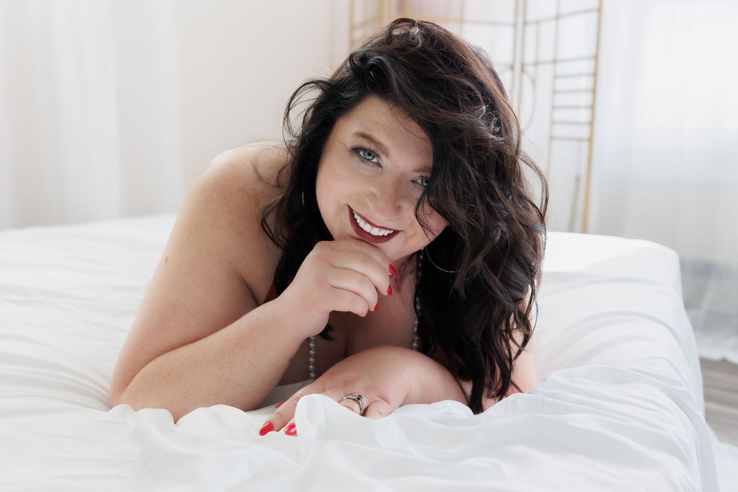 Bride in white lace lingerie and pearl-embellished veil sitting gracefully on a bed in soft natural light.