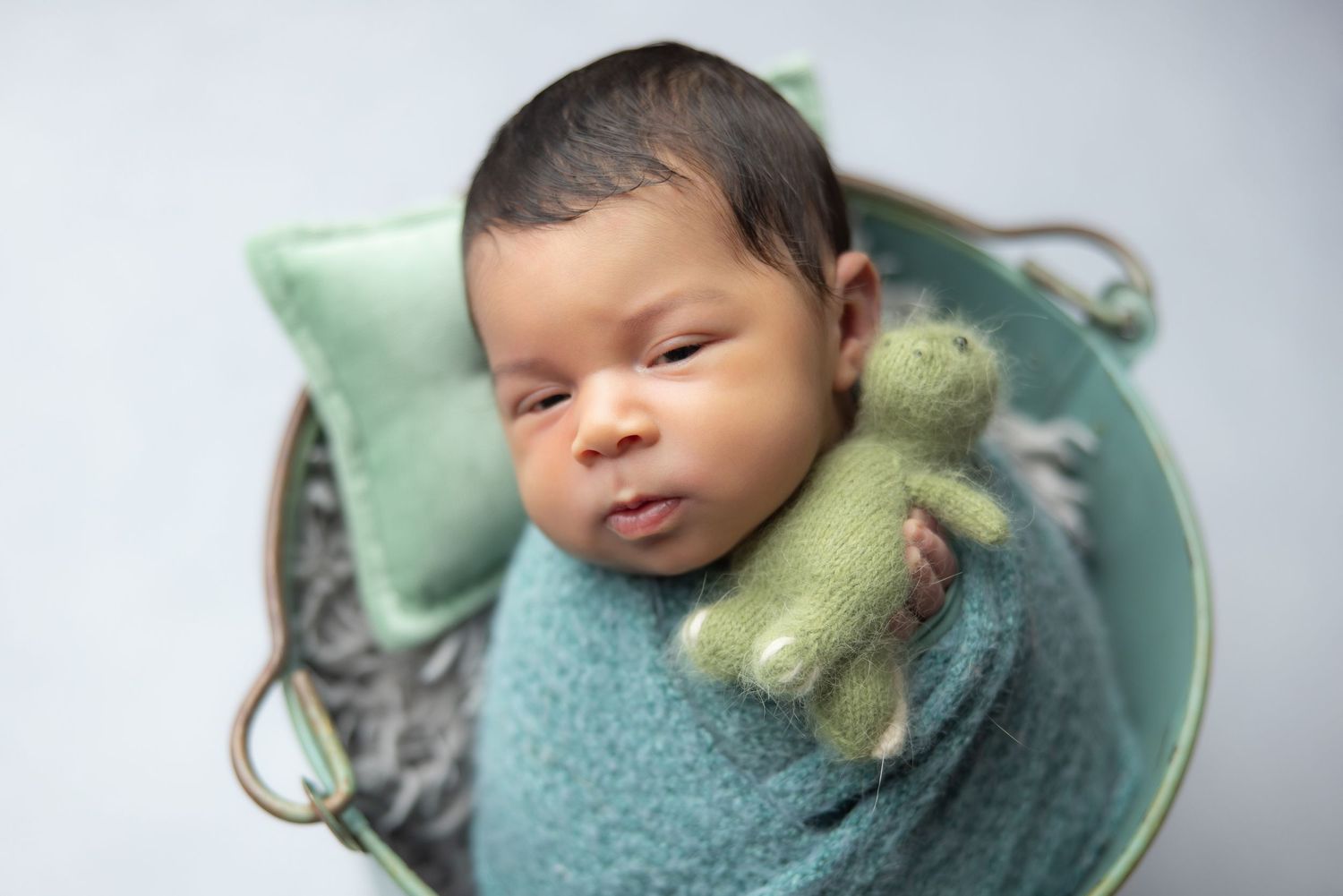 Newborn baby wrapped in teal swaddle holding green knit bear toy, resting in decorative bucket.