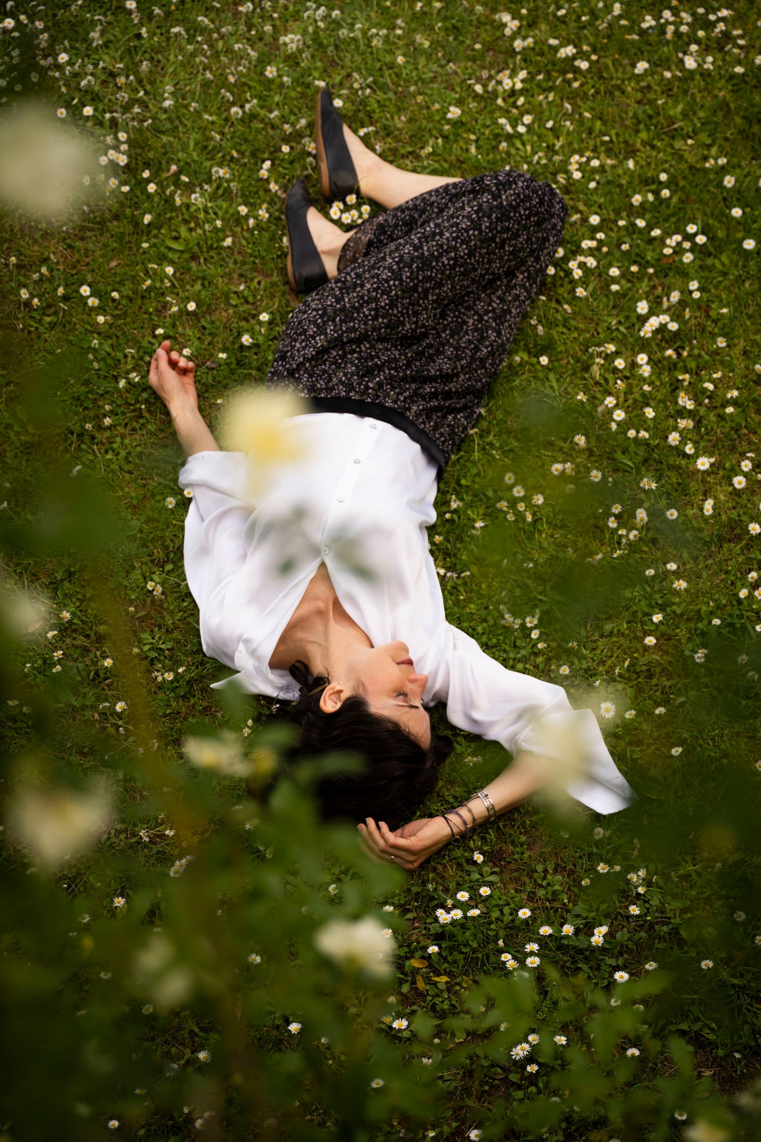 Woman in white blouse and floral skirt lying peacefully in a green field of white daisies, viewed from above.